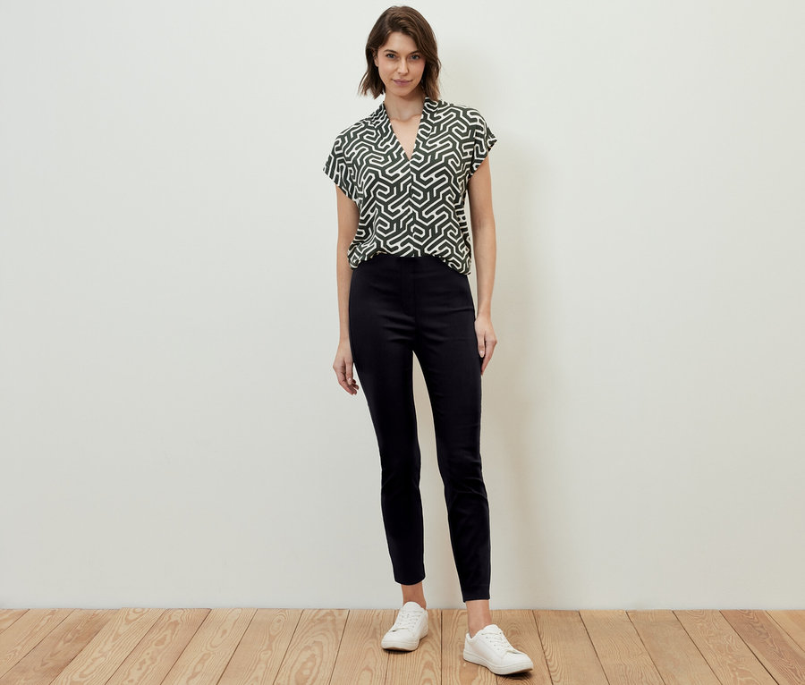 Une femme debout porte un pantalon stretch noir et un chemisier à motifs.