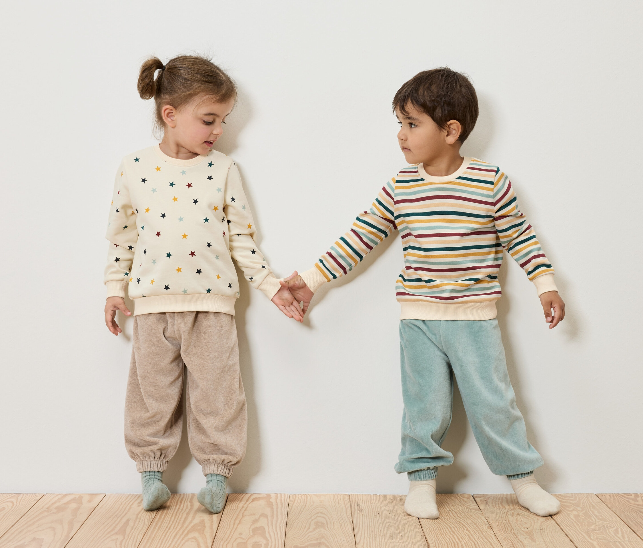 Deux enfants se tiennent la main. La fille porte un sweat-shirt crème avec des étoiles et un pantalon beige. Le garçon porte un sweat-shirt rayé et un pantalon bleu.