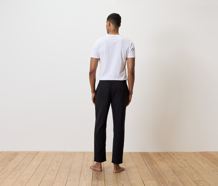 Un homme se tient dos à la caméra, portant un t-shirt blanc et un pantalon noir rayé.