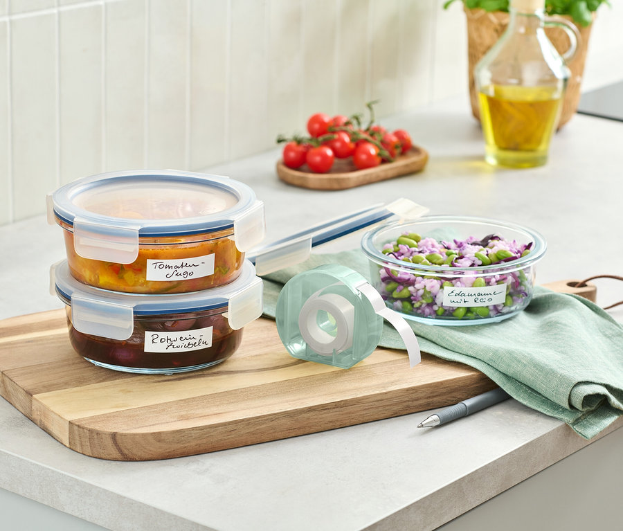 Deux récipients en verre remplis de nourriture empilés sur une planche à découper en bois à côté d'un rouleau d'étiquettes.