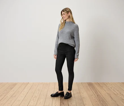 Une femme se tient sur un plancher en bois et porte un pull gris, un pantalon noir et des chaussures noires.