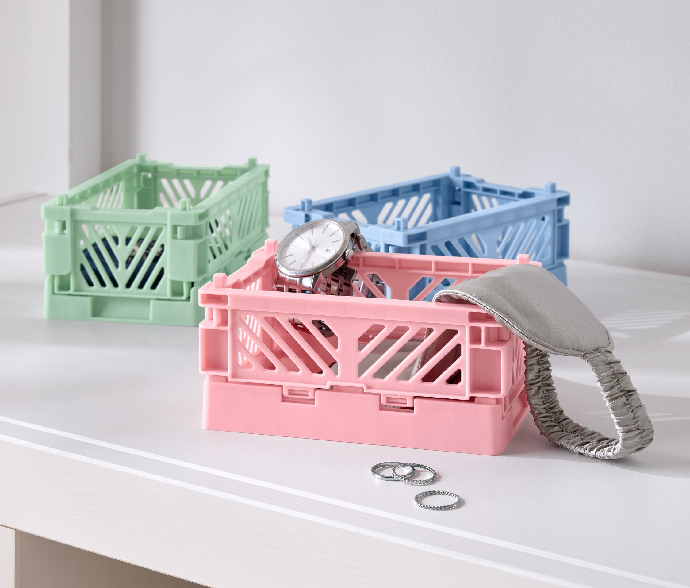 Trois boîtes pliantes colorées sur une table, une rose avec une montre, une bleue et une verte.