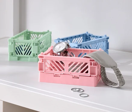 Trois boîtes pliantes colorées sur une table, une rose avec une montre, une bleue et une verte.