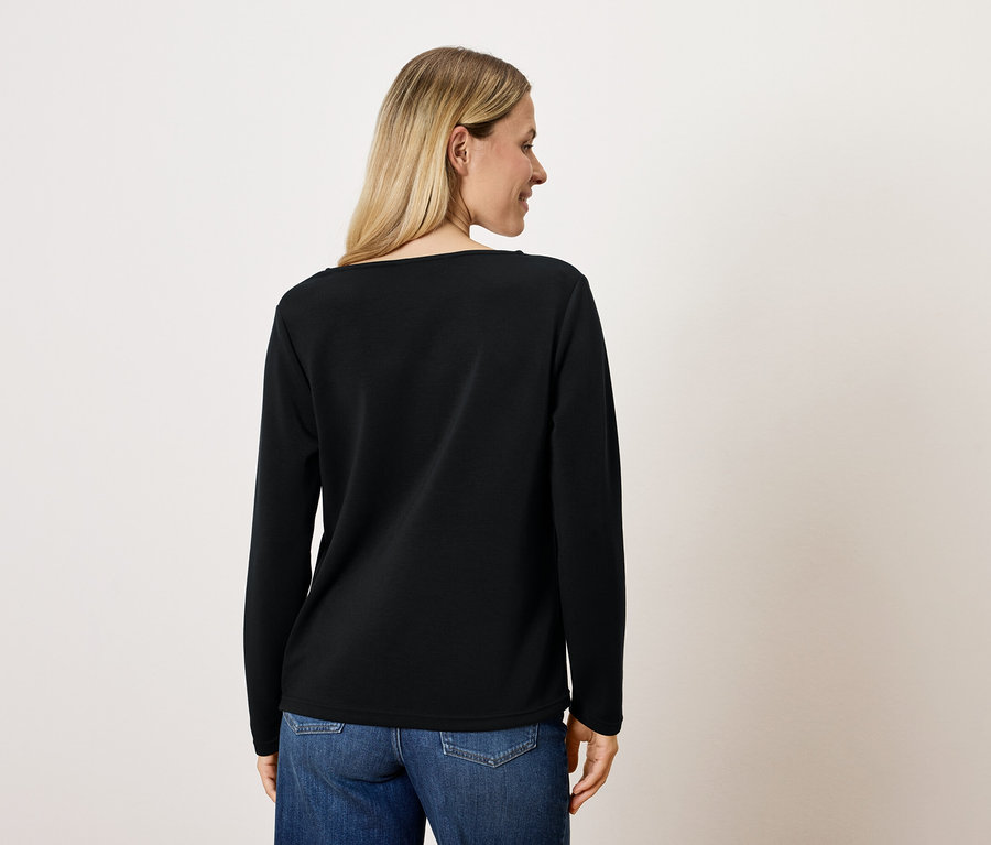 Une femme blonde, vue de dos, porte un T-shirt noir à manches longues et un jean bleu.