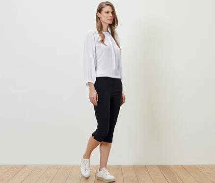 Une femme debout sur un plancher en bois porte un chemisier blanc et un pantalon 3/4 en bengaline noir.