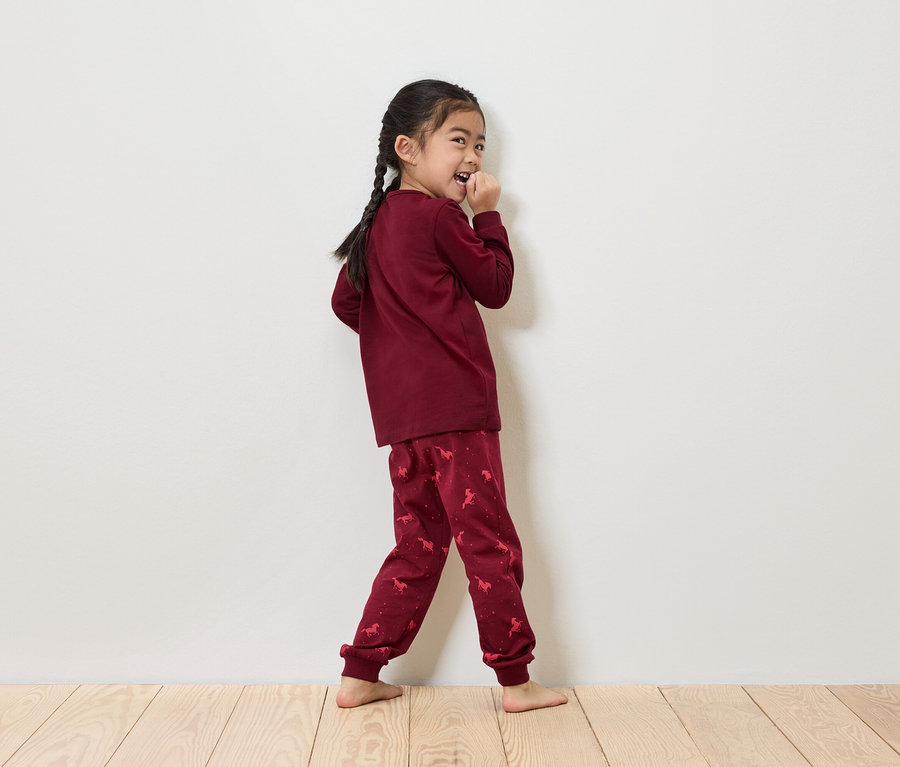 Fillette avec des tresses en pyjama bordeaux avec imprimé de licorne debout devant un mur blanc.