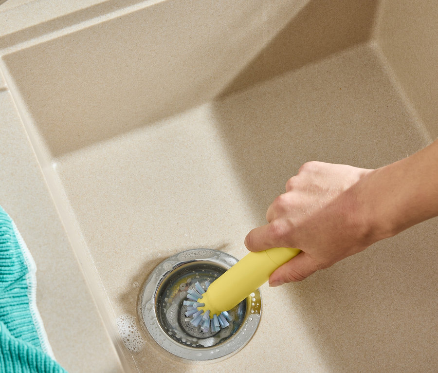 Une main nettoie le drain d'un évier avec une brosse jaune. Un torchon turquoise est visible à gauche.