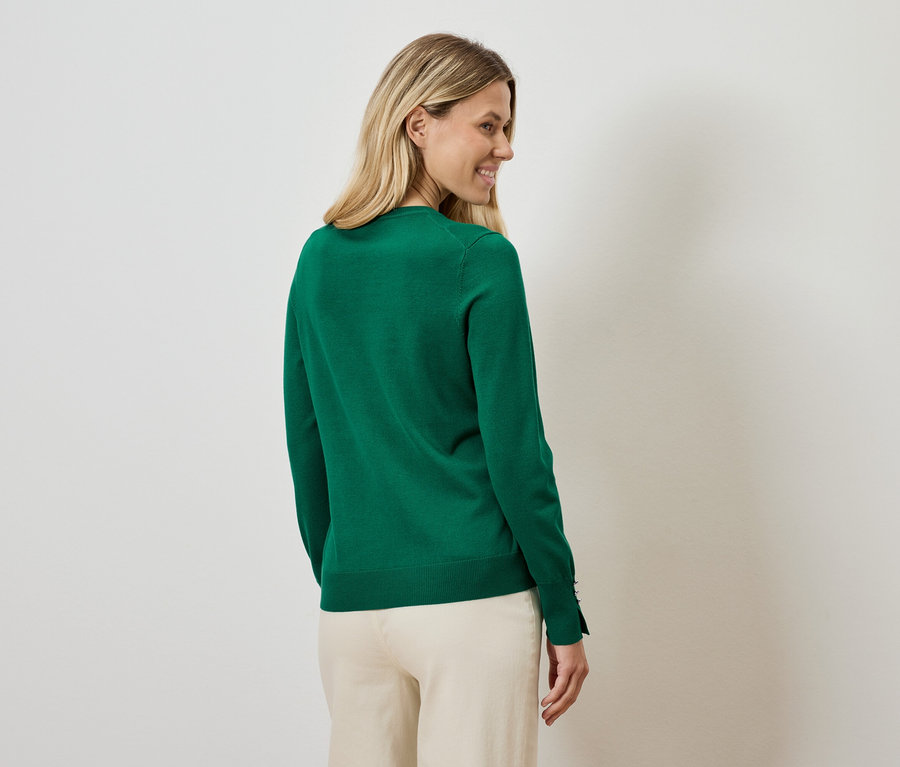 Une femme regarde par-dessus son épaule et porte un pull vert et un pantalon beige.