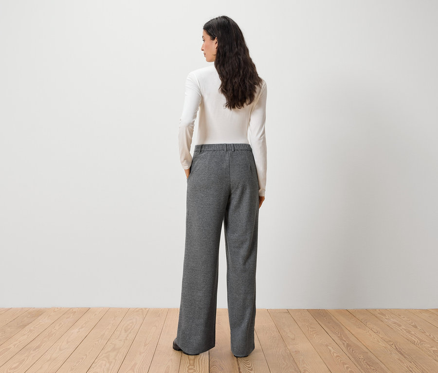 Une femme, dos à la caméra, porte un haut blanc à manches longues et un pantalon gris.