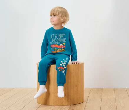 Un garçon est assis sur un tabouret en bois et porte un survêtement bleu avec un imprimé Hot Wheels.