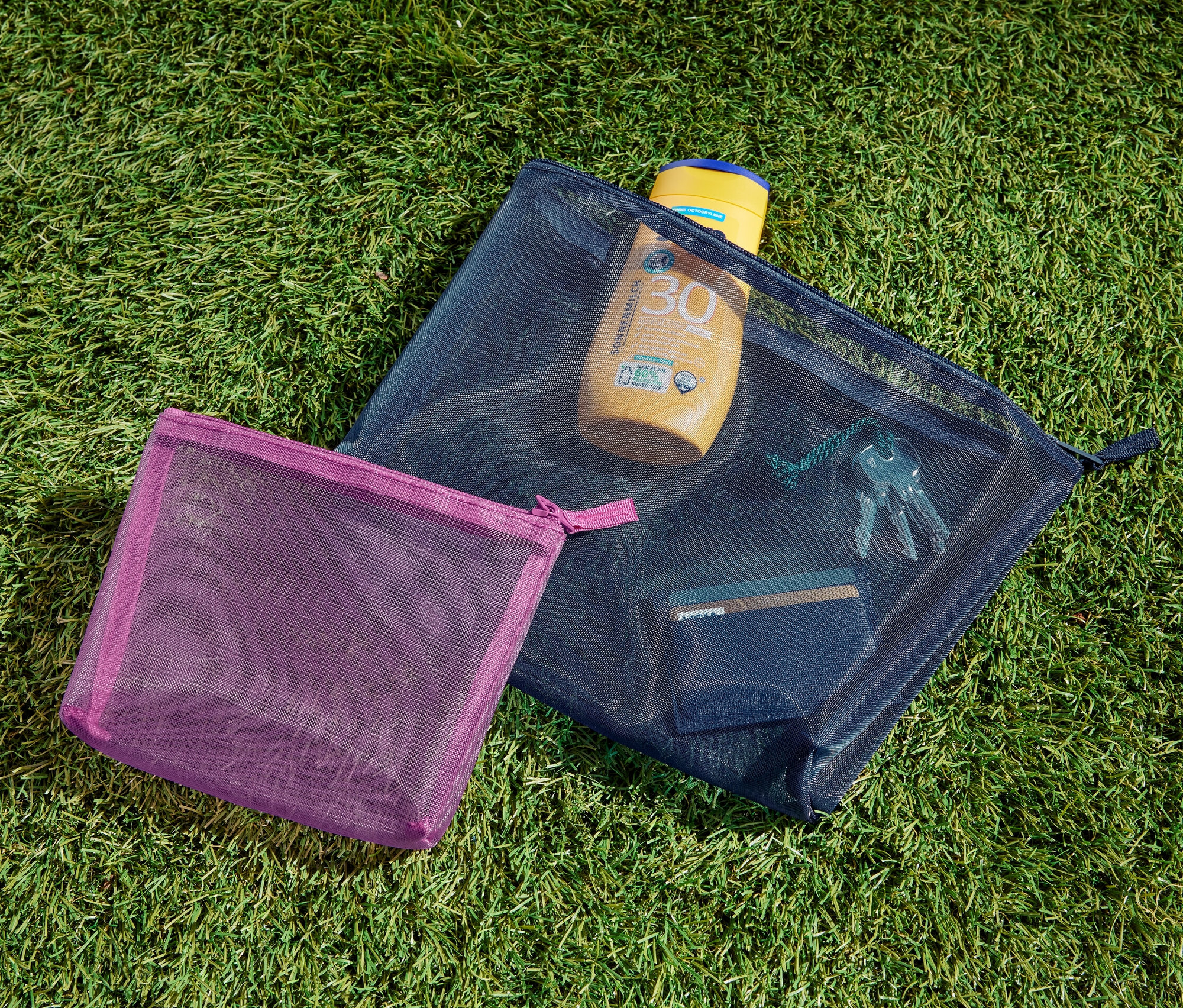 Deux pochettes de rangement sont posées sur l'herbe. L'une contient de la crème solaire, des clés et une carte.