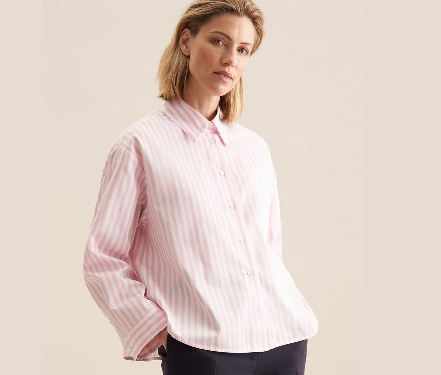 Une femme porte une chemise oversize rose rayée Seidensticker et un pantalon bleu marine.
