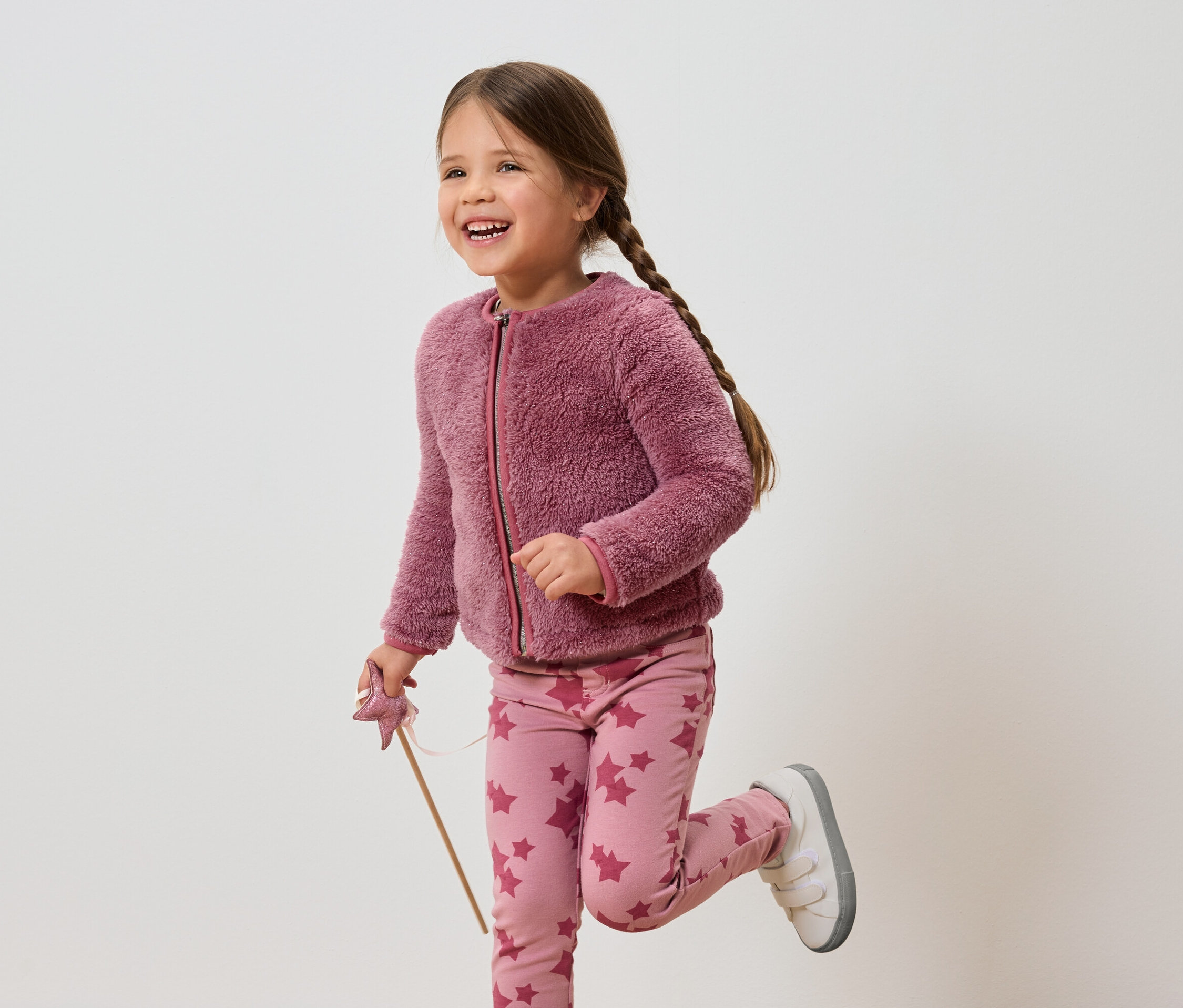 Fillette souriante aux cheveux bruns, veste rose et pantalon à étoiles, tenant une baguette.