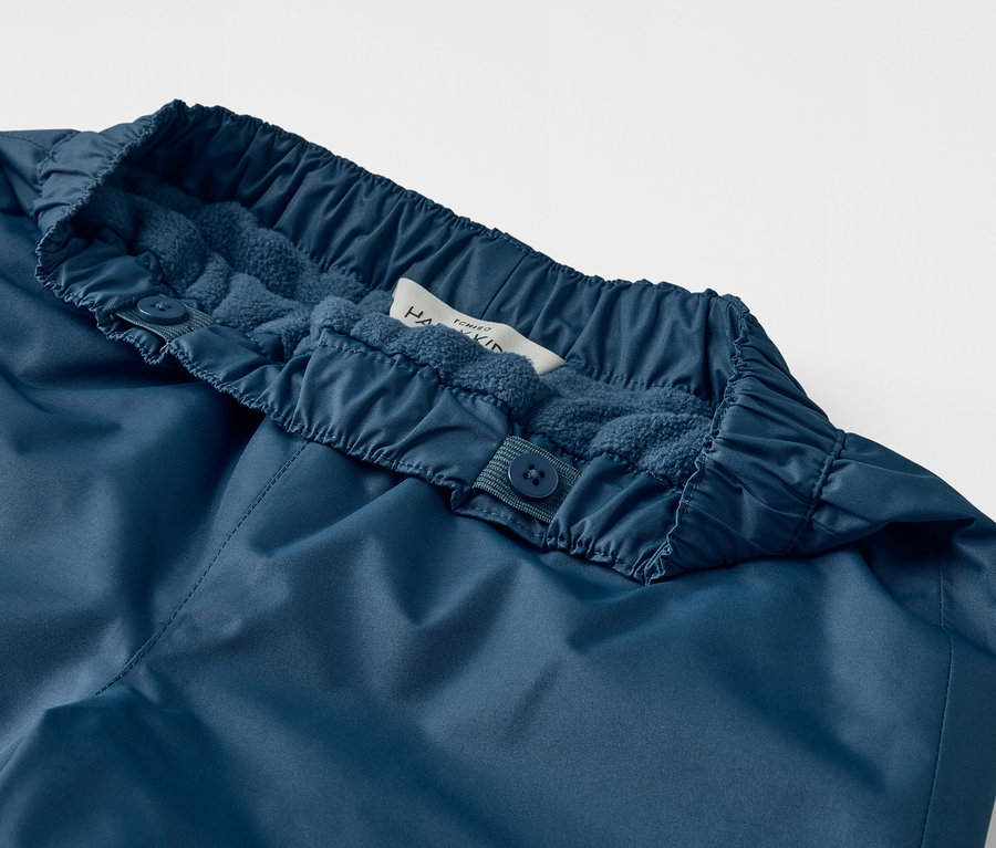 Gros plan sur un pantalon de pluie évolutif pour enfant, bleu, avec des boutons à la taille.
