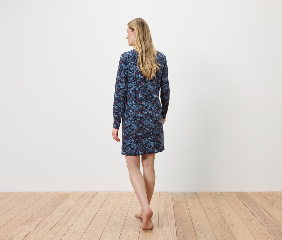 Une femme, dos à la caméra, marche pieds nus sur un plancher en bois, portant une robe bleue fleurie à manches longues.