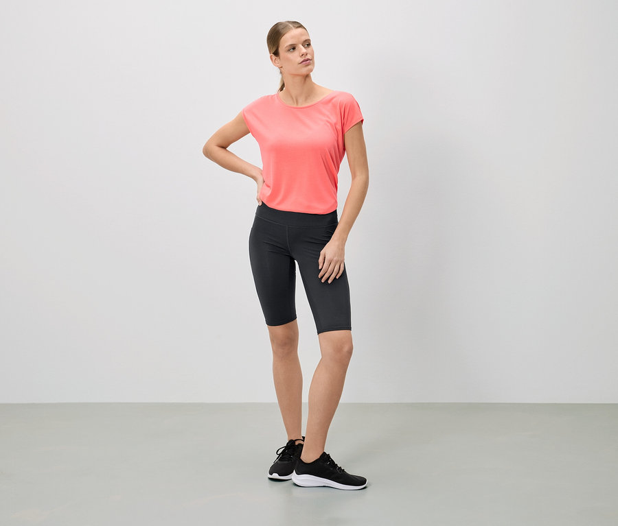 Une femme se tient debout en t-shirt rose, short de cycliste noir et baskets.