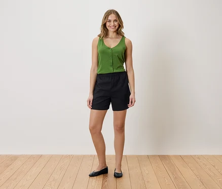 Femme portant un débardeur vert et un short noir, debout sur un plancher en bois devant un mur blanc.