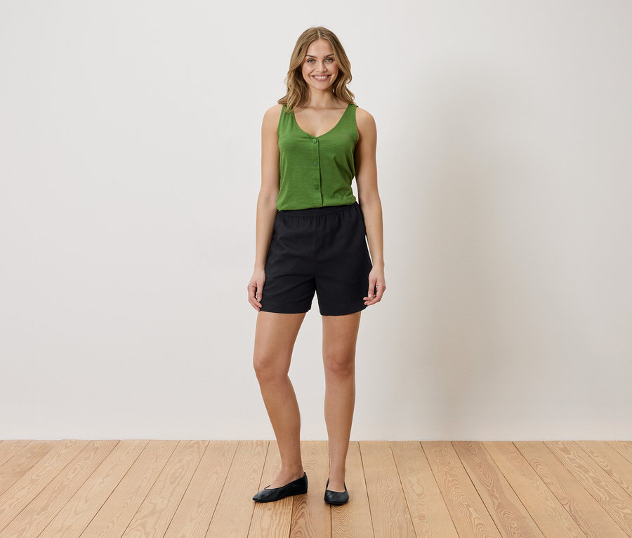 Femme portant un débardeur vert et un short noir, debout sur un plancher en bois devant un mur blanc.