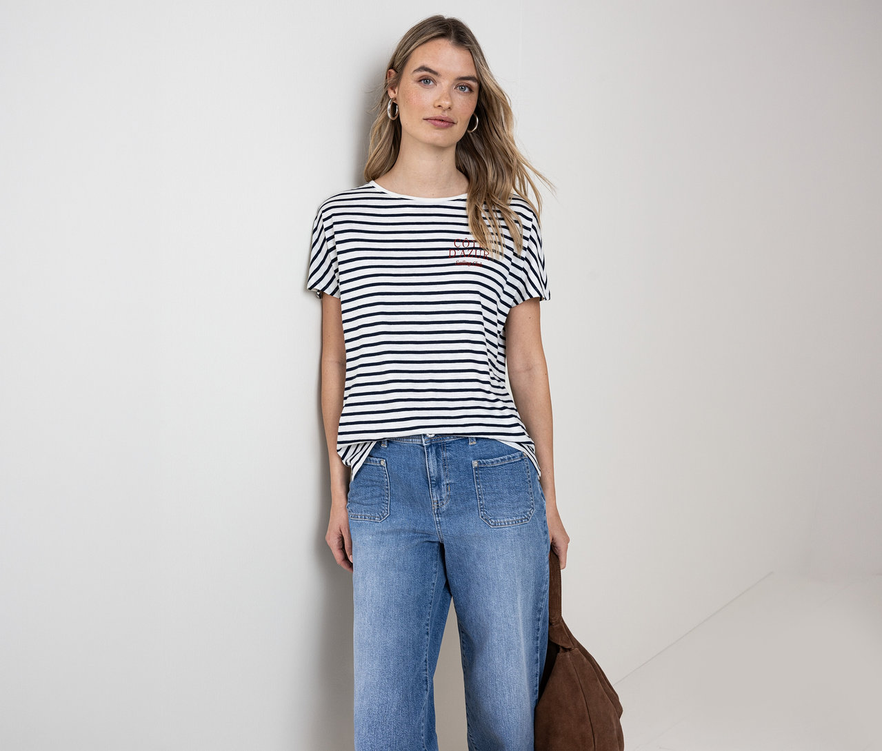 Une femme porte un t-shirt Street One bleu marine et blanc, un jean bleu et tient un sac marron.