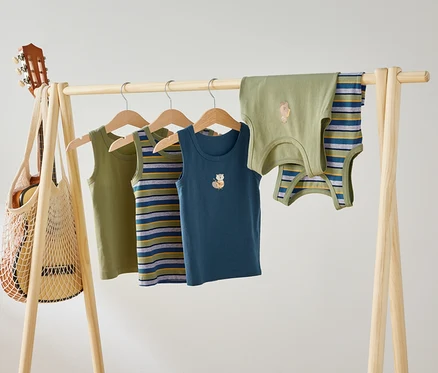 Quatre débardeurs pour enfants, vert, rayé et bleu avec des motifs d'animaux, sont suspendus à une tringle en bois. Une guitare est suspendue à la tringle dans un sac en filet.