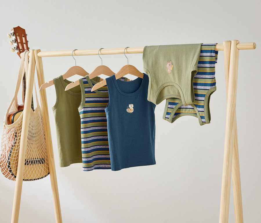 Quatre débardeurs pour enfants, vert, rayé et bleu avec des motifs d'animaux, sont suspendus à une tringle en bois. Une guitare est suspendue à la tringle dans un sac en filet.