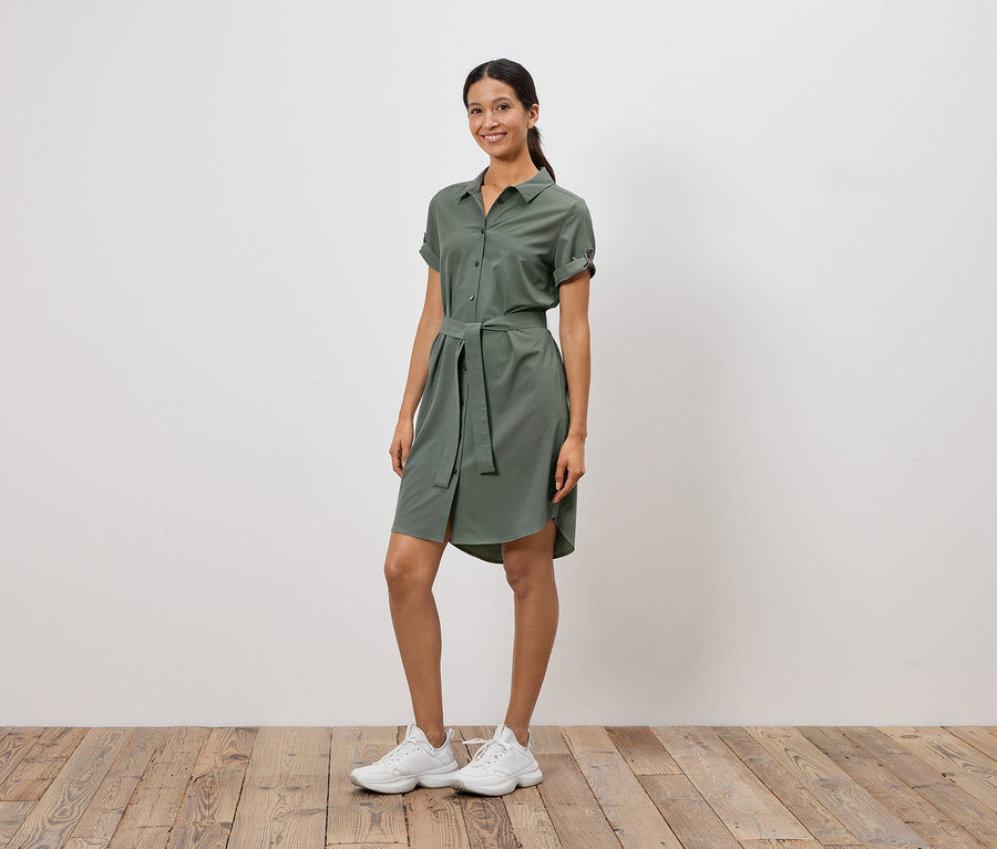 Une femme se tient devant un mur blanc, portant une robe chemise vert olive avec une ceinture et des baskets blanches.