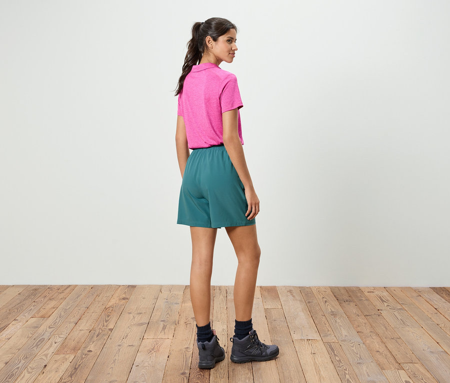 Une femme se tient dos à la caméra, elle porte un polo rose et un short vert.