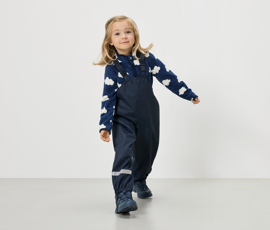 Une fillette blonde porte une veste en polaire bleu marine à motif de nuages et un pantalon à bretelles bleu marine.