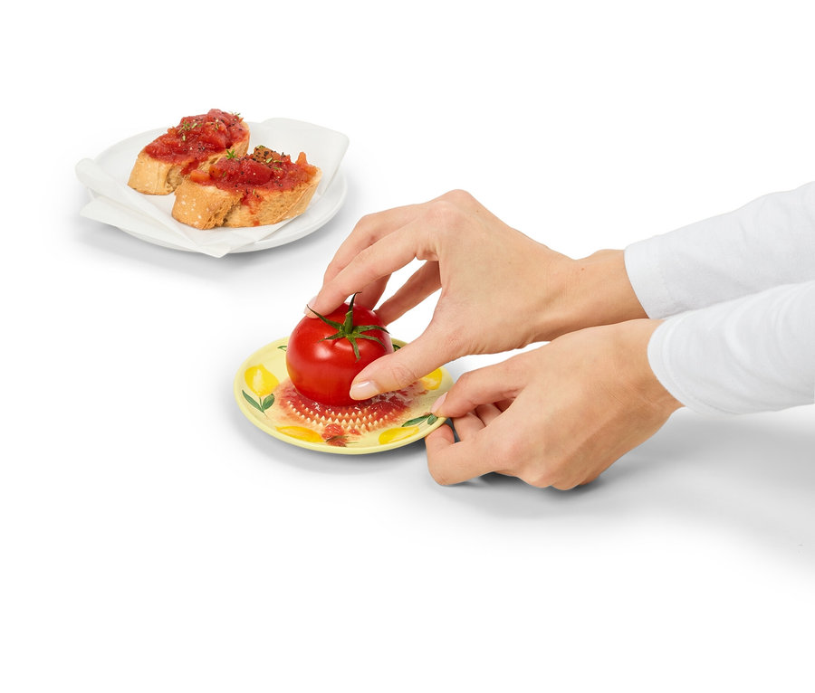 Des mains tiennent une assiette jaune avec une tomate rouge. En arrière-plan, deux pains à la tomate sur une assiette blanche.