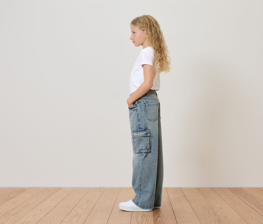 Une jeune fille aux cheveux blonds bouclés se tient de profil, portant un t-shirt blanc, un jean ample avec une poche latérale et des baskets blanches.