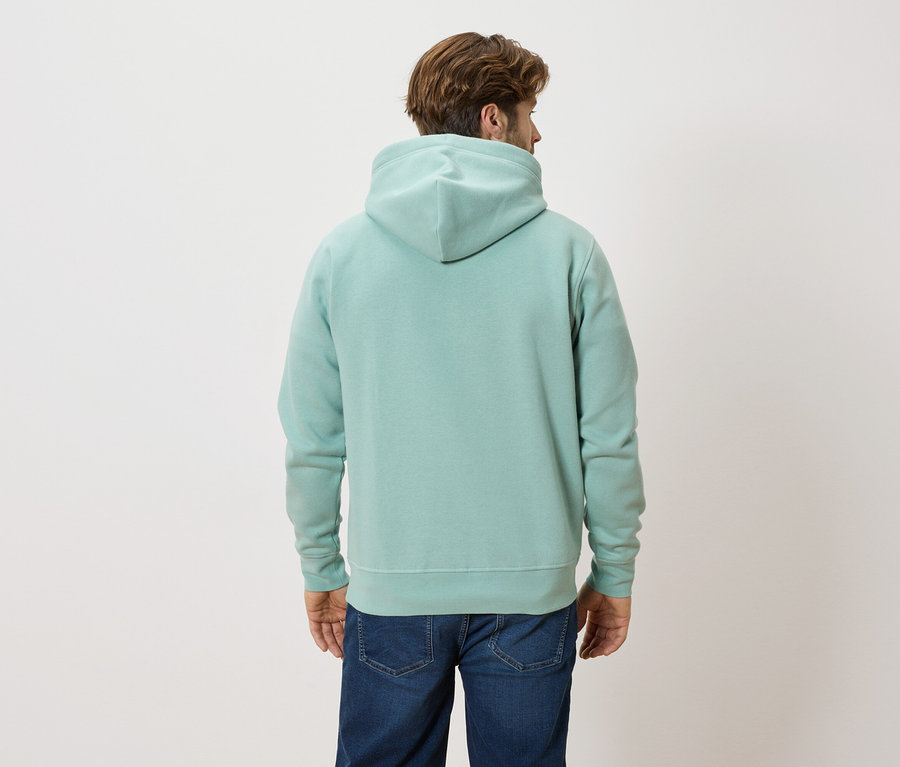 Un homme aux cheveux bruns porte un sweat à capuche vert menthe et un jean bleu.
