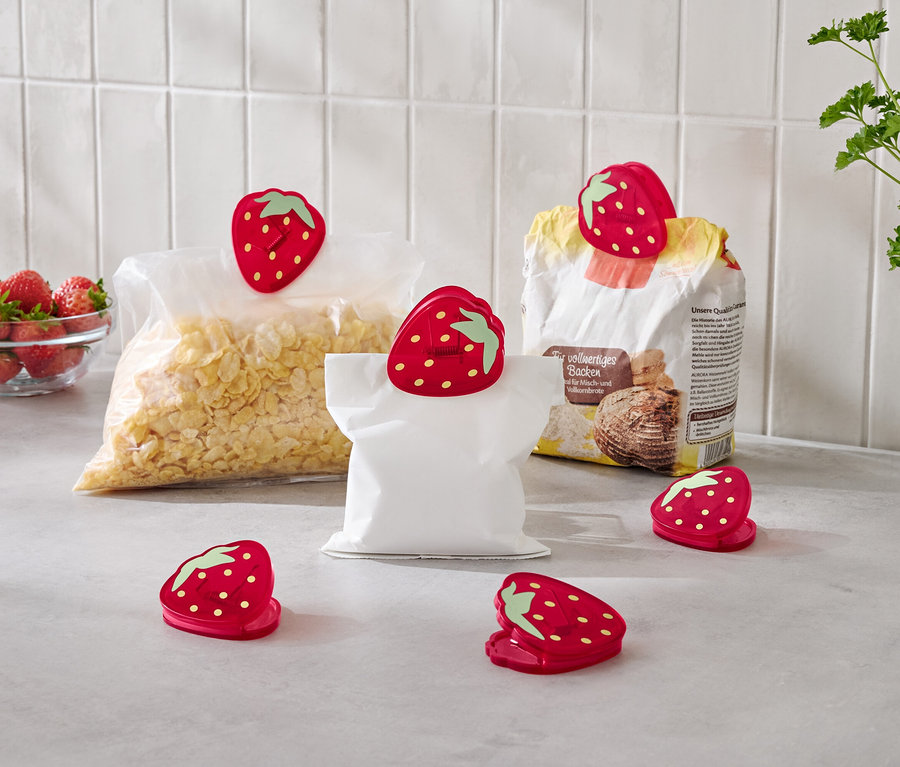 Sur un comptoir de cuisine, un bol de fraises, un sac de flocons de maïs, un sac de farine et un sac vide sont tous fermés par des clips rouges en forme de fraise.