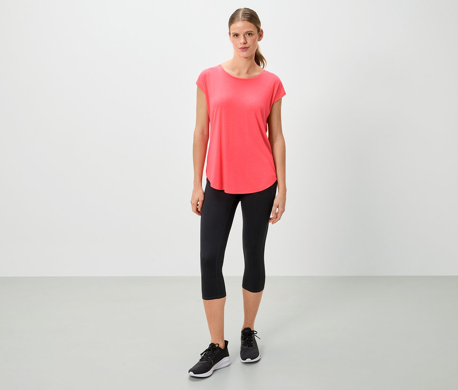 Femme portant un t-shirt corail, un legging noir et des baskets debout sur un fond blanc.
