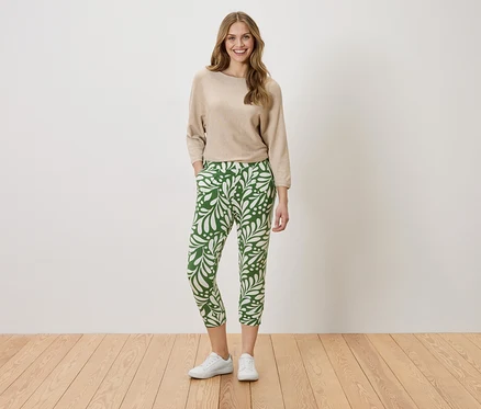 Une femme se tient debout, les mains dans les poches, devant un mur. Elle porte un pull beige et un pantalon vert avec un motif de feuilles blanches.