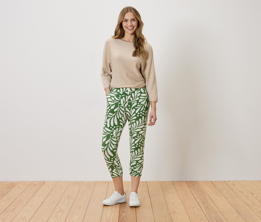 Une femme se tient debout, les mains dans les poches, devant un mur. Elle porte un pull beige et un pantalon vert avec un motif de feuilles blanches.
