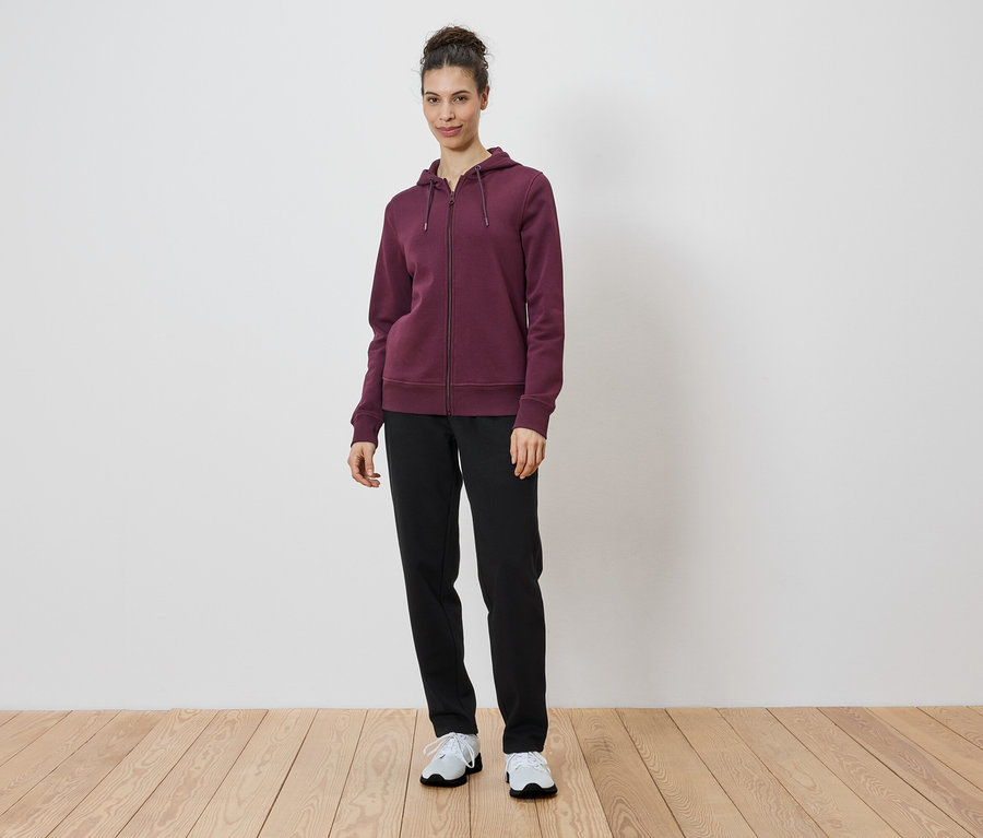 Femme debout portant un sweat à capuche bordeaux zippé, un pantalon noir et des baskets blanches.