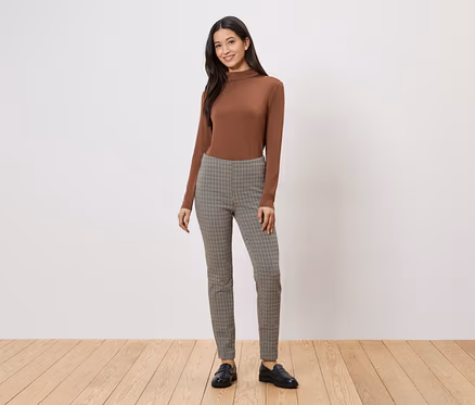 Une femme portant un pull à col roulé marron et un pantalon à carreaux pose sur un plancher en bois devant un mur blanc.