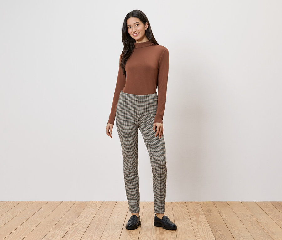 Une femme portant un pull à col roulé marron et un pantalon à carreaux pose sur un plancher en bois devant un mur blanc.