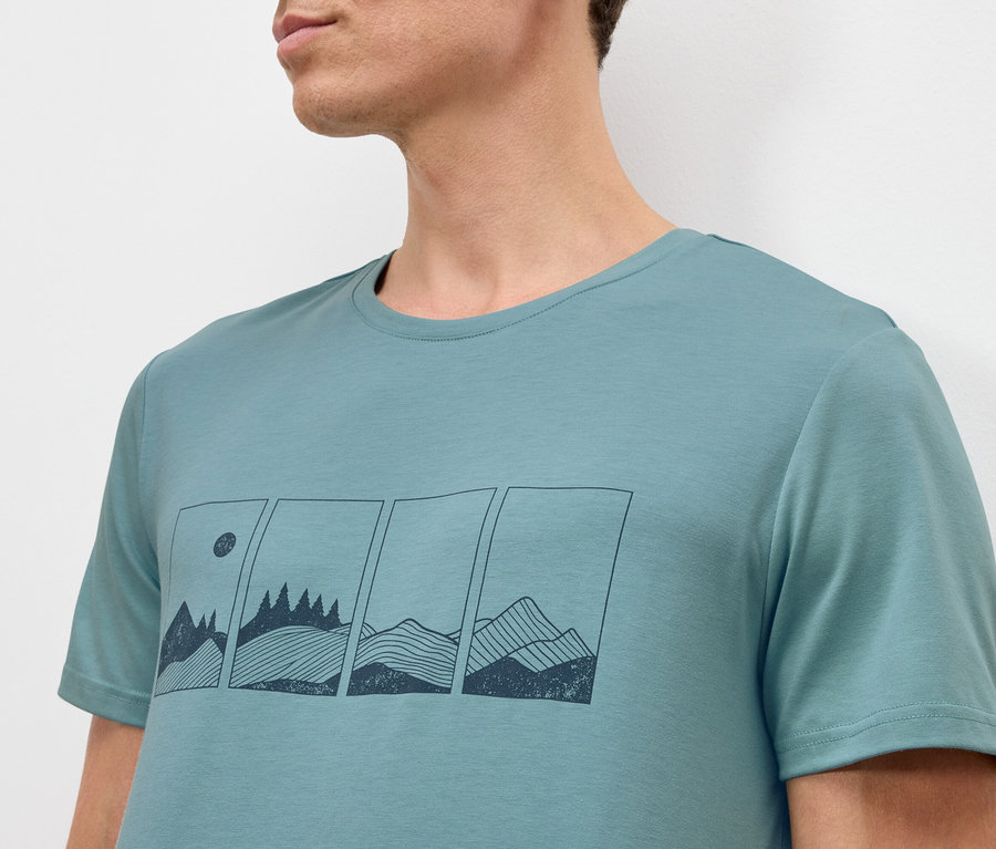 Un homme porte un t-shirt turquoise avec un graphisme de montagnes et d'arbres dans des rectangles.
