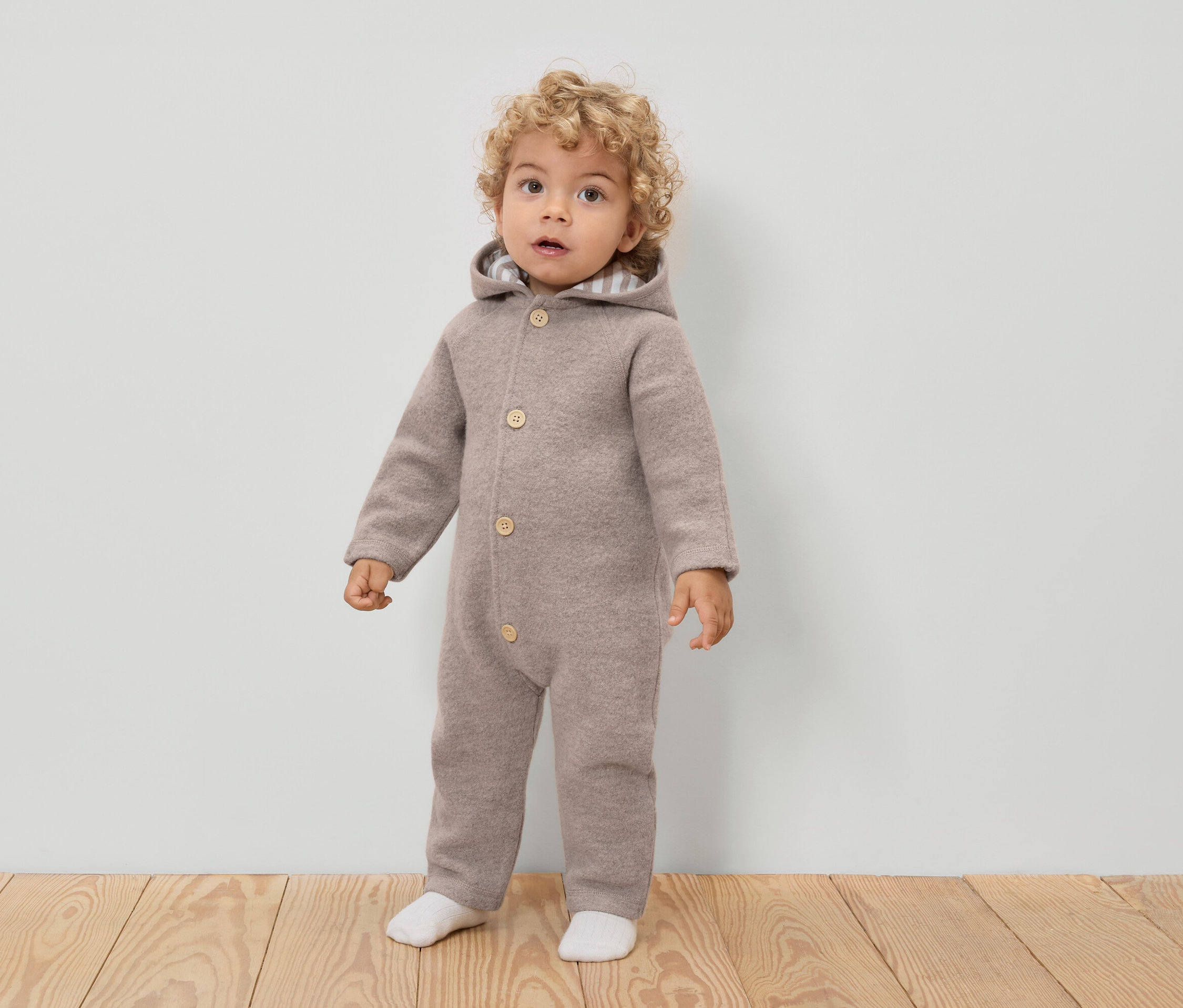 Un jeune enfant aux cheveux bouclés porte une combinaison en laine marron clair à capuche et à boutons, ainsi que des chaussettes blanches.