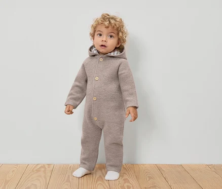 Un jeune enfant aux cheveux bouclés porte une combinaison en laine marron clair à capuche et à boutons, ainsi que des chaussettes blanches.