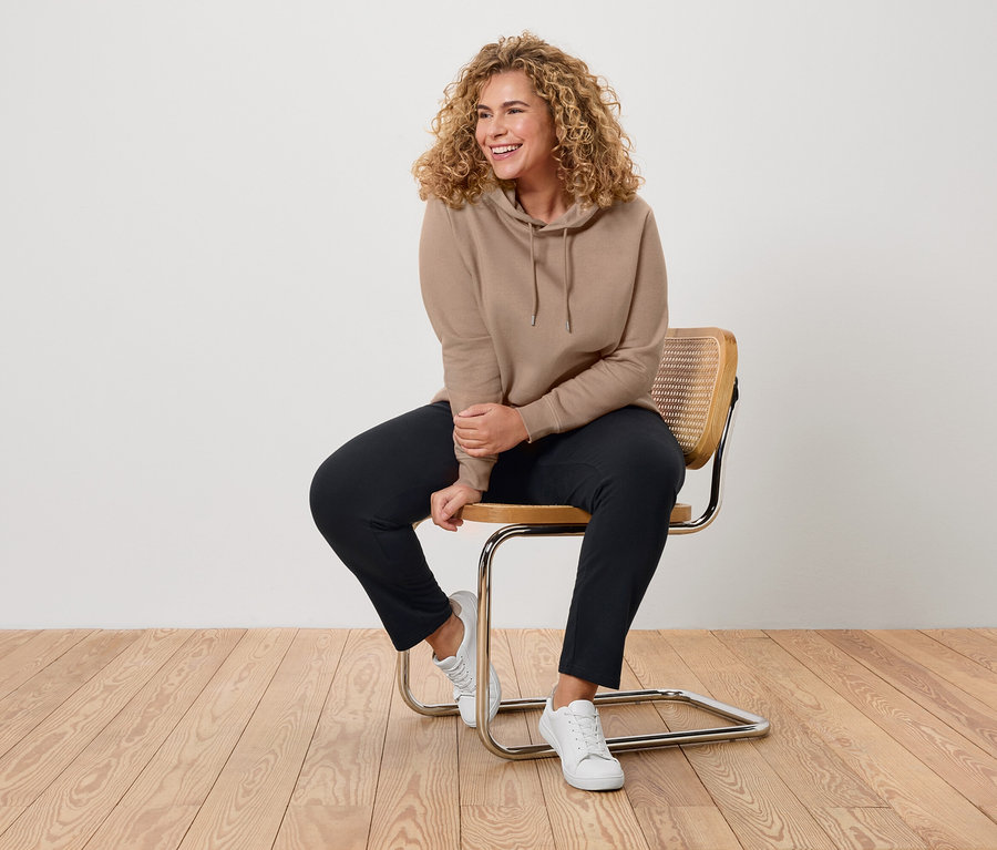 Une femme aux cheveux bouclés est assise sur une chaise, portant un sweat à capuche beige, un pantalon noir et des baskets blanches.