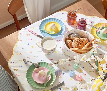 Table à manger avec couverts, comprenant des assiettes avec des œufs, des tasses, des couverts, une corbeille à pain, des pots de confiture, des décorations de Pâques et un chemin de table fleuri.