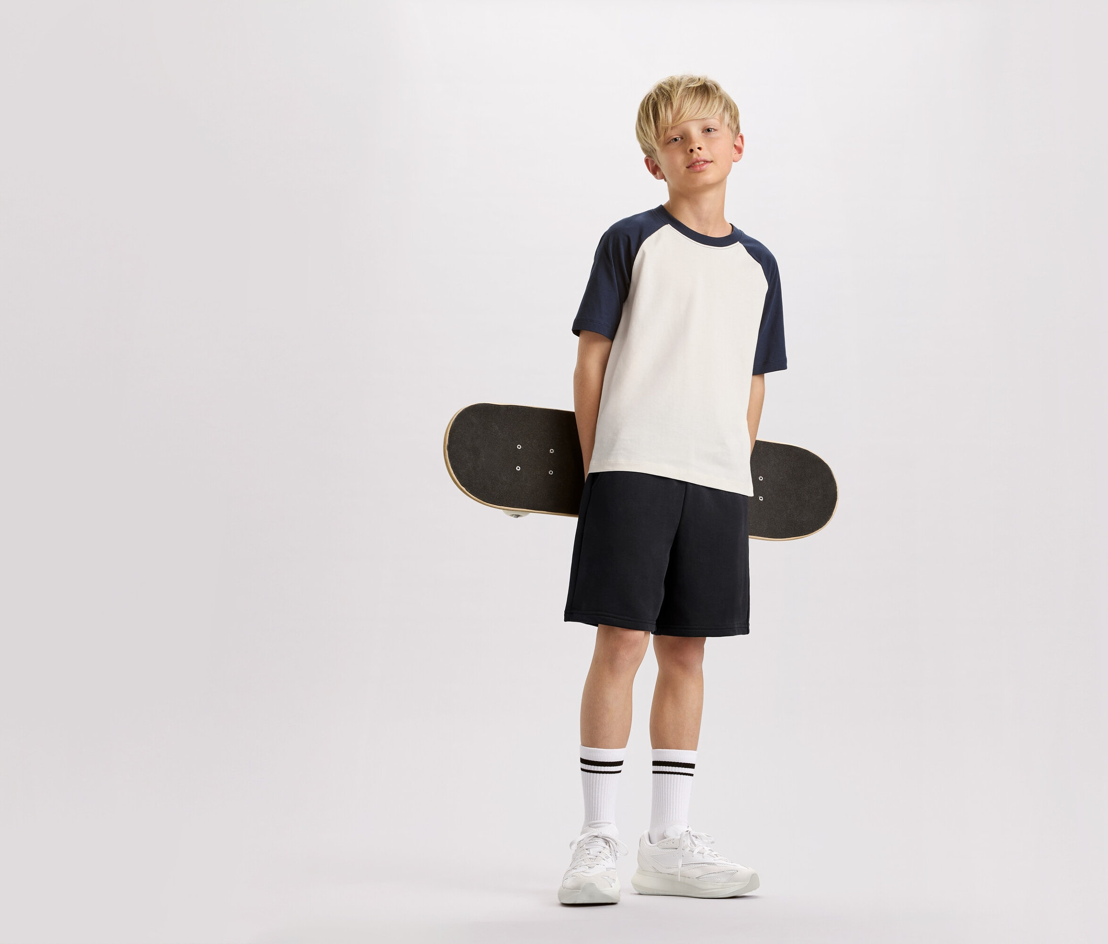 Un garçon blond tient un skateboard dans son dos, vêtu d'un t-shirt à manches bleu marine, d'un short et de baskets.