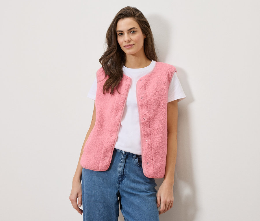 Une femme porte un gilet sans manches en peluche teddy rose sur un t-shirt blanc et un jean bleu.