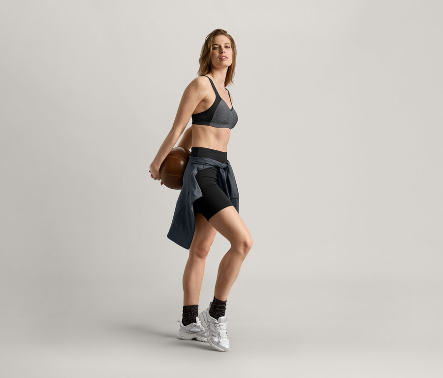 Femme tenant un ballon de basket dans le dos, portant un soutien-gorge de sport, un short et une veste de sport anthracite.