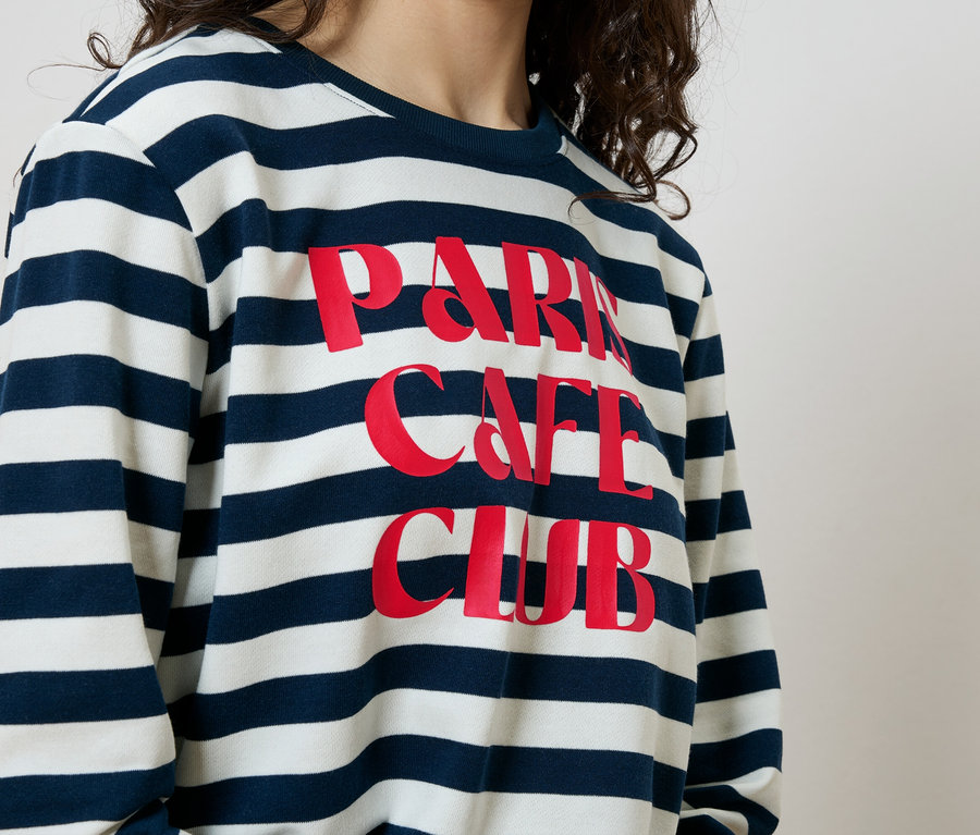Une femme porte un sweat-shirt rayé bleu et blanc avec l'inscription "Paris Cafe Club" en rouge.