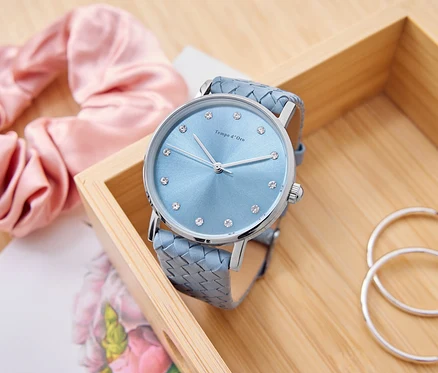 Une montre bleu clair avec des strass est posée dans une boîte en bois, à côté d'un chouchou rose, d'un bloc-notes et de créoles argentées.