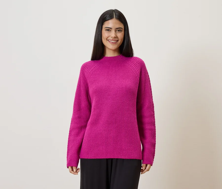Une femme aux cheveux foncés porte un pull en maille rose et un pantalon noir sur un fond clair.