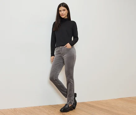 Une femme portant un pull à col roulé noir, un pantalon en velours côtelé gris et des mocassins noirs, debout devant un mur blanc.